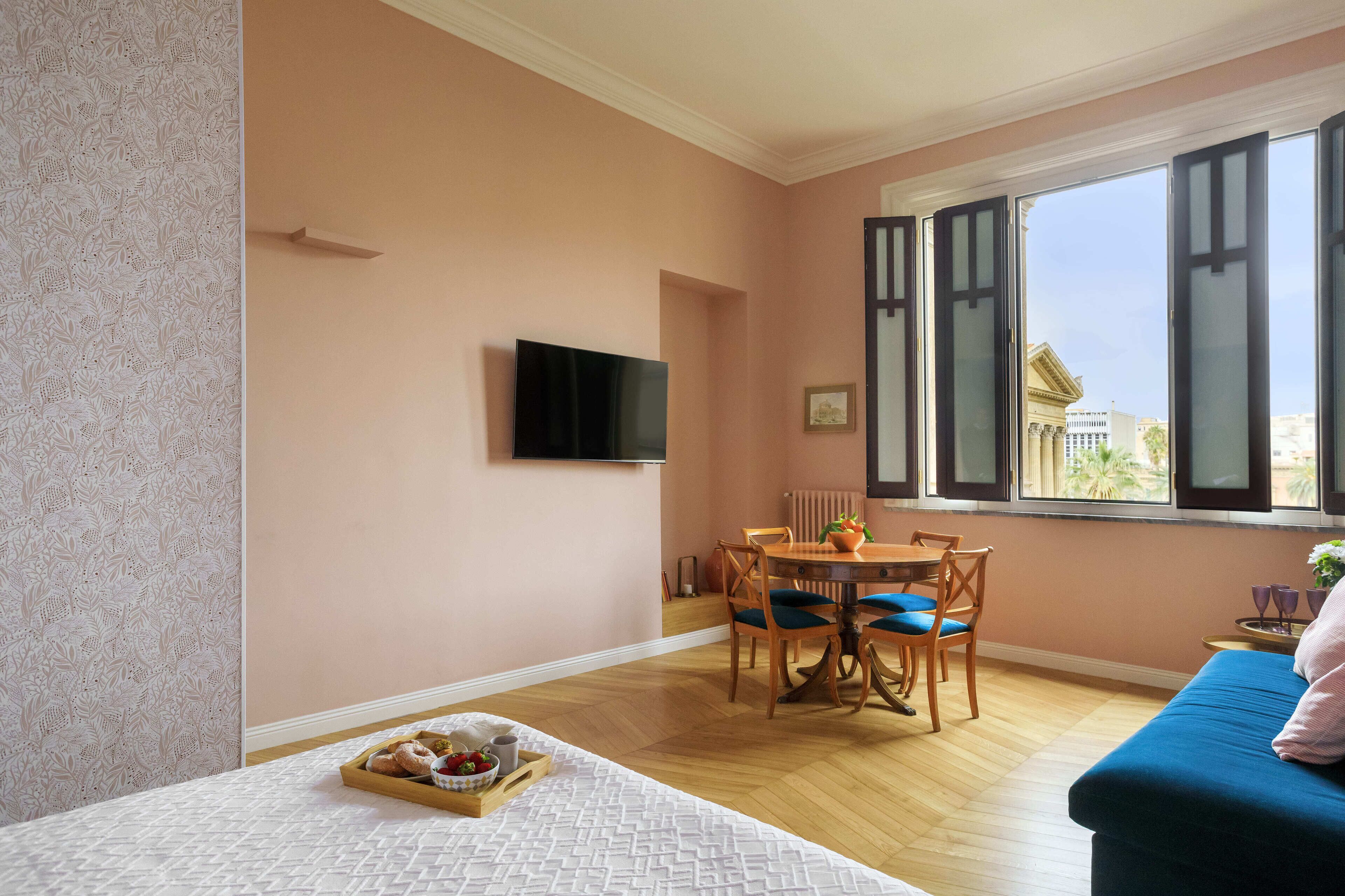 Una habitación con una cama, una mesa de comedor con sillas, un televisor montado en la pared y una ventana con persianas.
