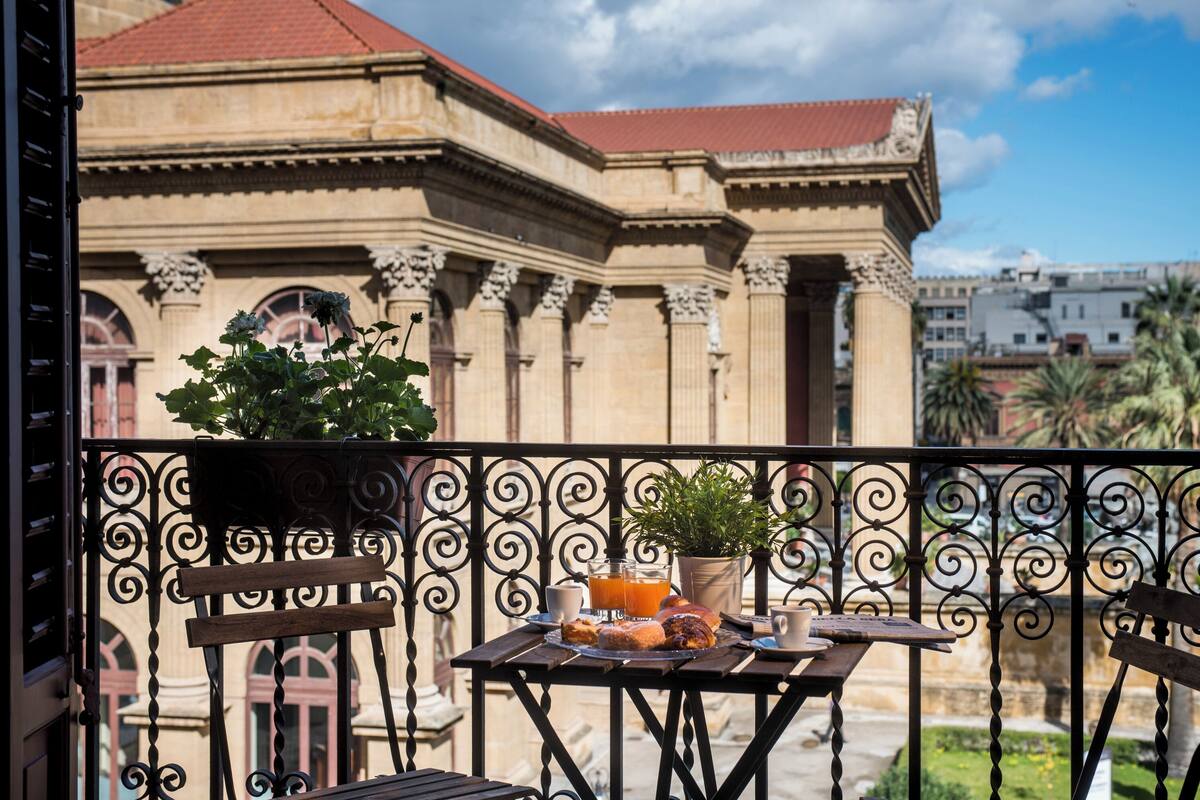 Un balcon avec une table préparée pour le petit-déjeuner, une plante en pot et une vue sur un bâtiment historique doté de colonnes et d’un toit rouge.
