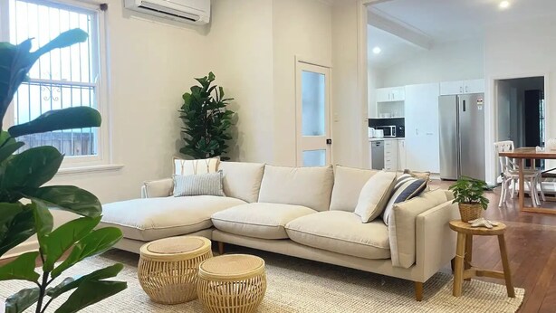 Ein modernes Wohnzimmer mit einem beigefarbenen Ecksofa, zwei Rattan-Beistelltischen, einer großen Topfpflanze und Blick in den Küchenbereich.
