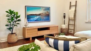 Ein Wohnzimmer mit einem Holz-Fernsehschrank, einem großen Gemälde einer Küstenlandschaft, einer Topfpflanze, einer Leiter und einem gestreiften Kissen.