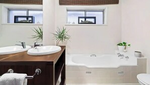 Ein modernes Badezimmer mit zwei Waschbecken, einer Badewanne und zwei Fenstern mit Jalousien.