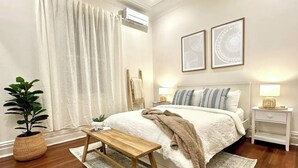 Ein Schlafzimmer mit einem Bett, Nachttischen, einer Bank, einer Topfpflanze und zwei gerahmten Bildern an der Wand.