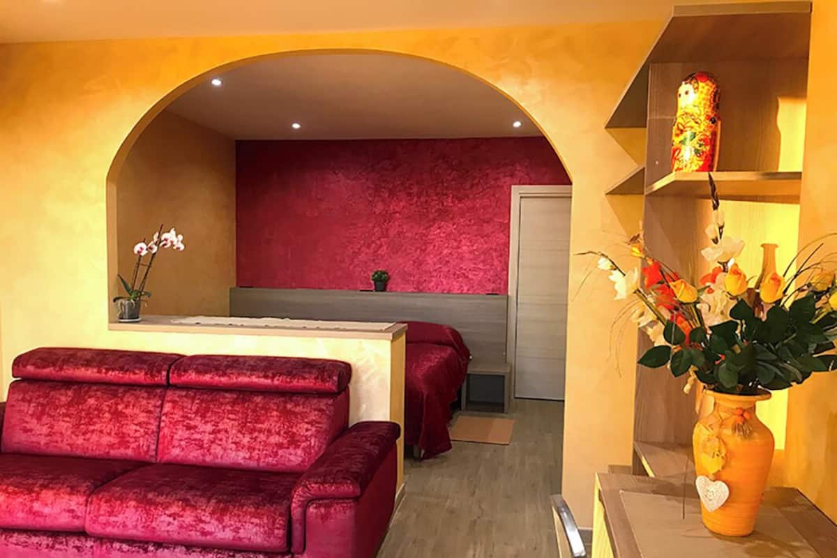 Un sofá de terciopelo rojo, una mesita y un jarrón con flores en una habitación con una pared de acento roja y un estante de madera.