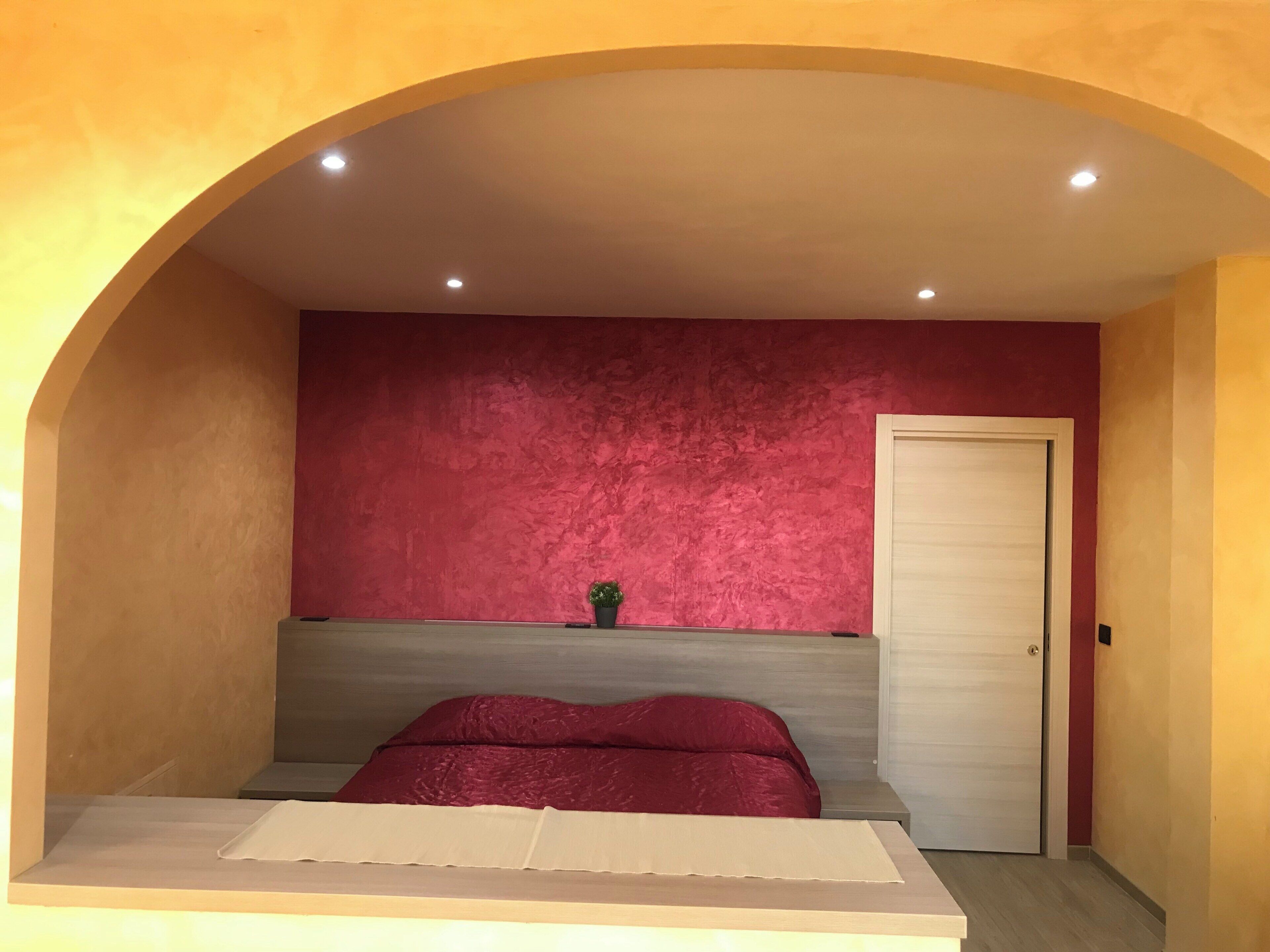 A bedroom with a bed, a small bedside table, a potted plant, and a door.