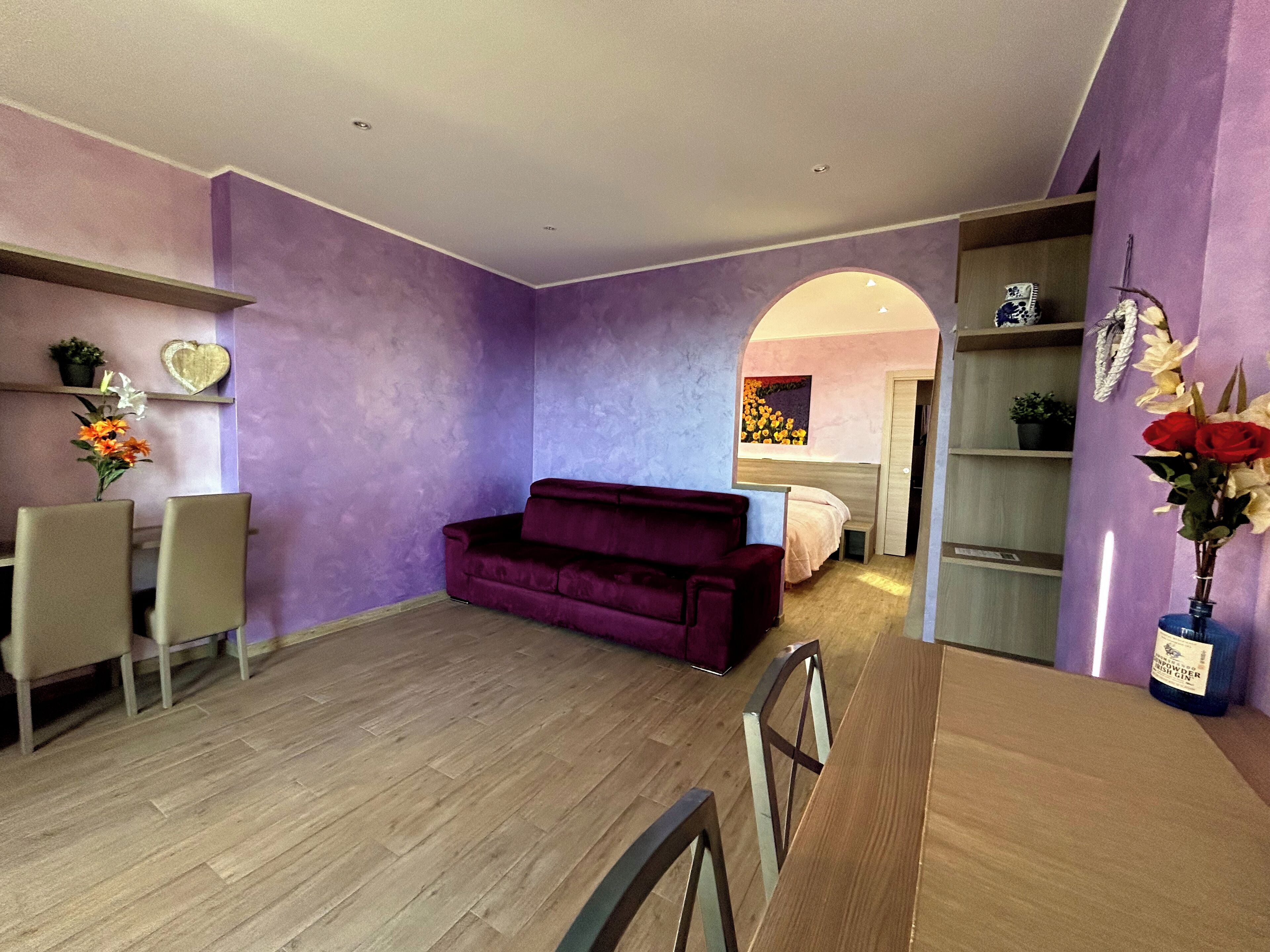 A modern living room with a purple wall, a wooden floor, a red sofa, and a dining area with a wooden table and chairs.