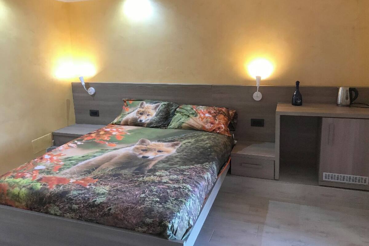 Un dormitorio con una cama con cubrecama con temática de zorro, lámparas de noche, una mesita de noche, un pequeño armario y una tetera sobre la mesita.