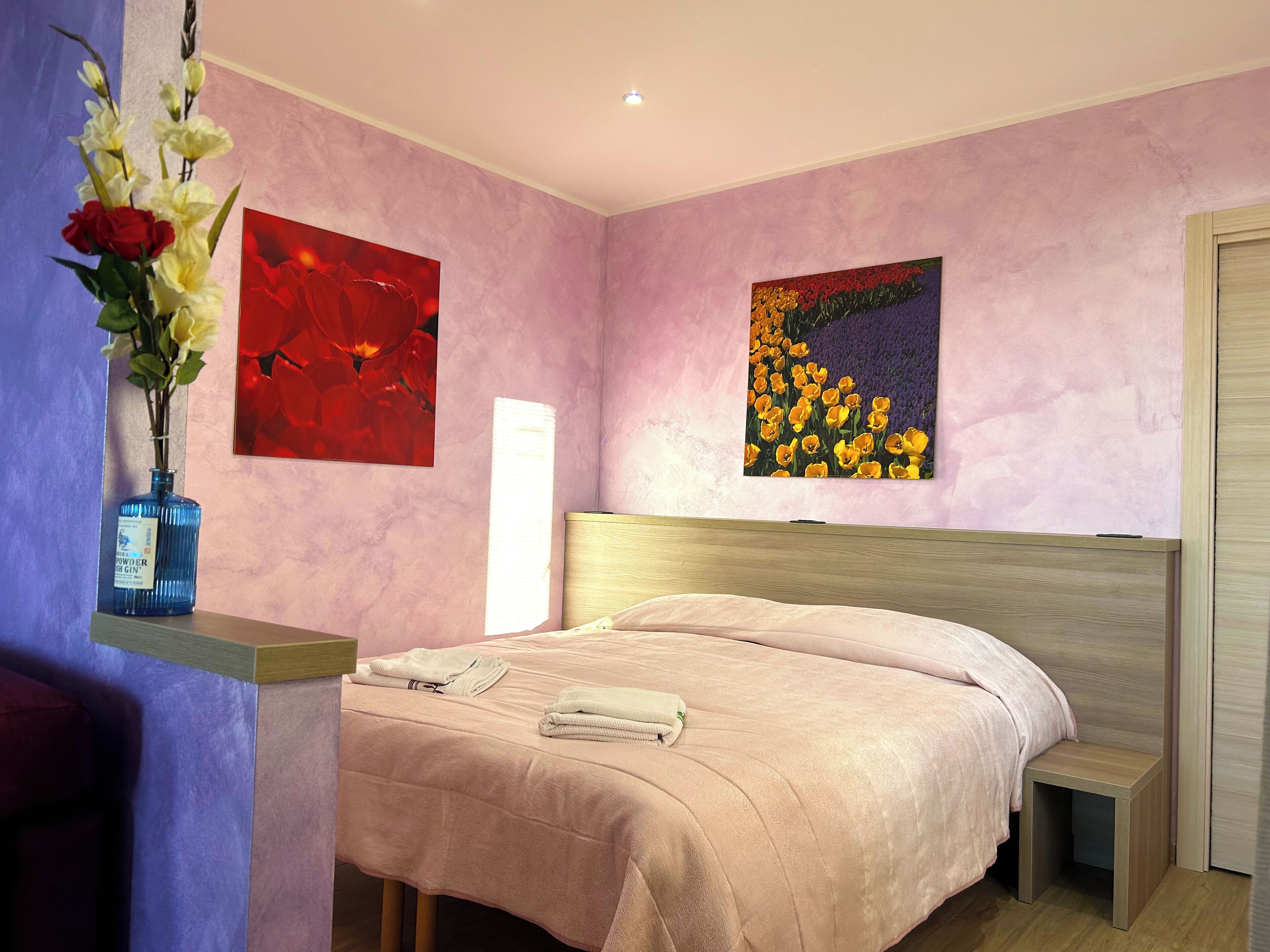 A bedroom with a bed, a nightstand, a vase with flowers, and two paintings on the wall.