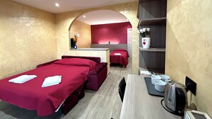 A hotel room with a bed, a desk, a chair, a small table, a shelf with a kettle, and a vase with flowers.