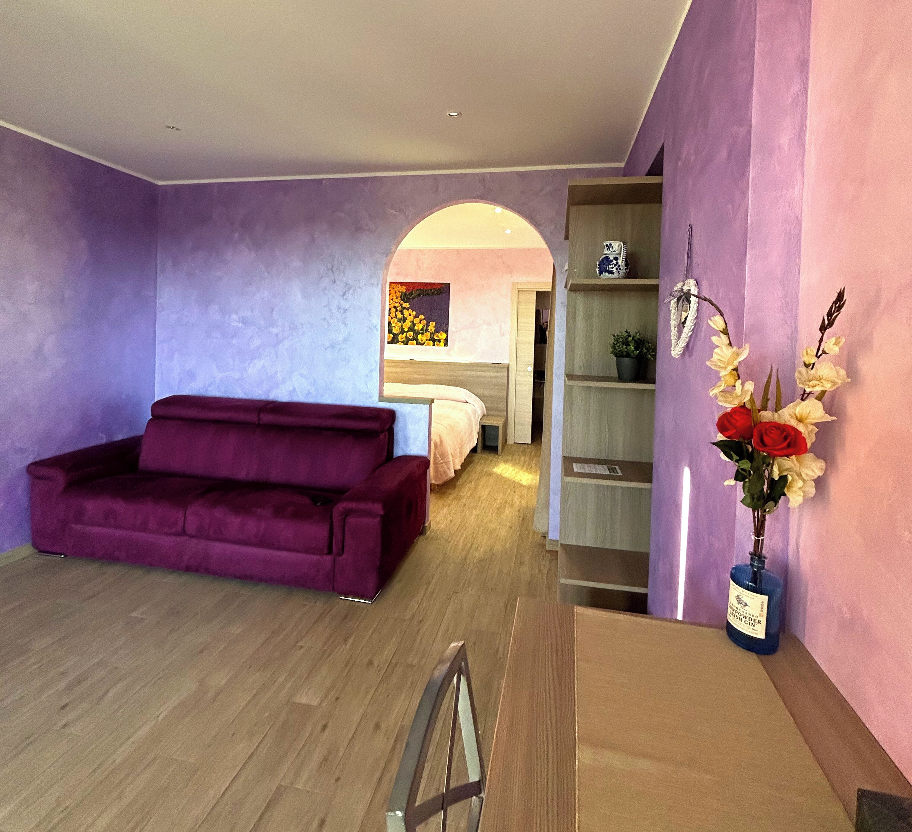 A room with a purple sofa, a bed, a wooden cabinet, and a vase with flowers.