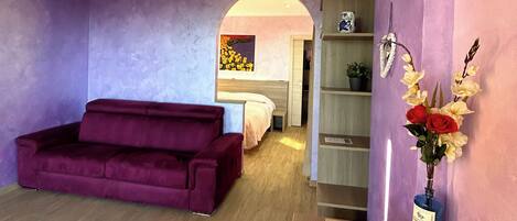 A room with a purple sofa, a bed, a wooden cabinet, and a vase with flowers.