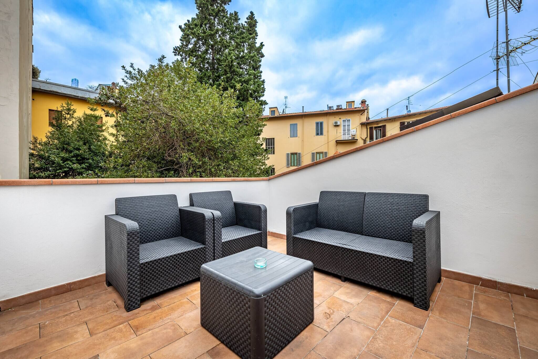 A rooftop patio with a dark wicker sofa set, a square ottoman, and a clear glass candle holder.