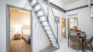 A modern interior with a staircase, a bed, a bedside table with a lamp, a dining area with a table and chairs, and an air conditioning unit. - YiD Donatello con terrazzo (Florence)