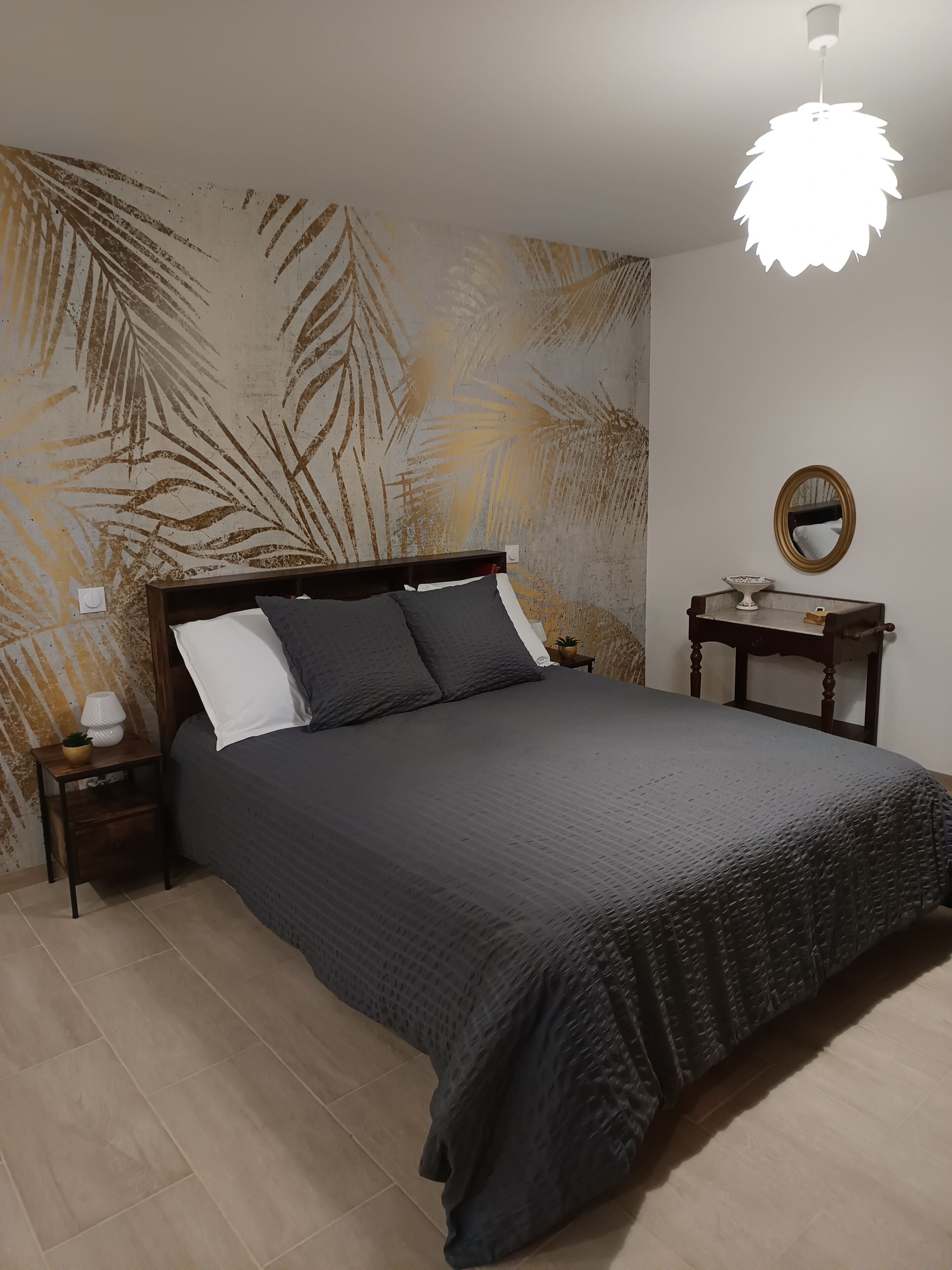 Une chambre à coucher comprenant un lit, des tables de chevet, une lampe, un miroir et un mur orné d’un motif de feuilles.