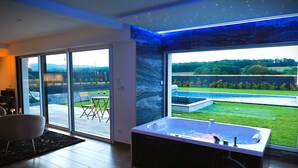 Une salle de bain moderne dotée d’une grande baignoire, d’une porte en verre donnant sur un espace extérieur, et offrant une vue sur une piscine et un jardin luxuriant.