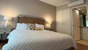 Una cama grande con edredón blanco acolchado, dos mesitas de noche con lámparas, un armario y un espejo.