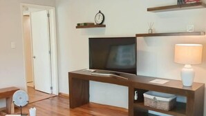 Una sala de estar con piso de madera, un televisor sobre un mueble de madera, una lámpara y una estantería.