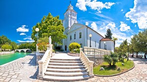 A historic building with a clock tower, a stone staircase leading to the entrance, and a paved courtyard with a small bridge over a body of water. - Pool House Beba Tribalj Lake (Vinodol)
