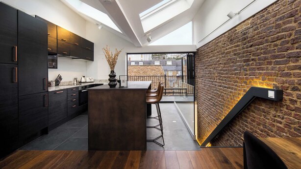 A modern kitchen with dark cabinetry, a central island, and a brick wall. - 1 Min Walk to Euston Station | Luxury Townhouse (London)