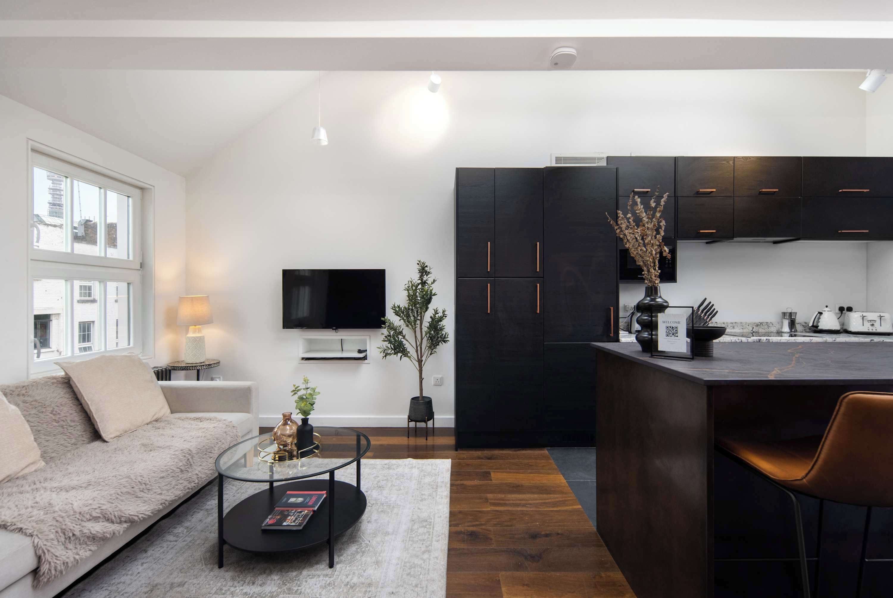 A modern living room with a sofa, coffee table, and a TV. There is a kitchen area with dark cabinets and a dining space with a chair.