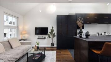 A modern living room with a sofa, coffee table, and a TV. There is a kitchen area with dark cabinets and a dining space with a chair.