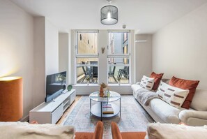 A modern living room with a flat-screen TV, a glass coffee table, a sofa with cushions, and large windows with a view of the outside. - 5 Mins Walk to Moorgate | Tranquil City Retreat (London)