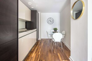 A modern kitchen with dark cabinetry, a wooden floor, a dining area with white chairs, a table, and a wall clock.