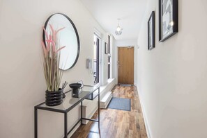 A hallway with a mirror, a vase with reeds, a black metal console table, a wooden door, and a light fixture.