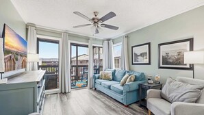 A spacious living room with a blue sofa, a grey sofa, a ceiling fan, and large windows offering a view of the outside. - Sea Blue Suite at Tilghman w Pool and Hot Tub (North Myrtle Beach)