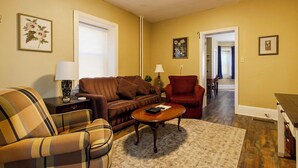 Un salon avec un canapé marron, un fauteuil à carreaux, une table basse en bois et un fauteuil rouge. On y trouve une lampe sur une table d’appoint, un tableau encadré au mur et une porte qui donne sur une autre pièce.