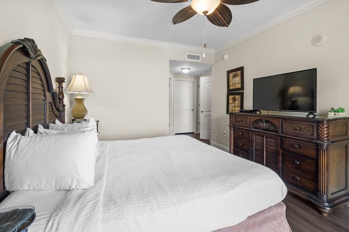 A bedroom with a large bed, a ceiling fan, a TV, a dresser, and a lamp.