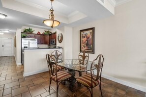 A dining area with a glass table and metal chairs, a kitchen in the background, and a ceiling light fixture. - Shoreline Splendor Sanctuary: 2BR Condo with WiFi (North Myrtle Beach)