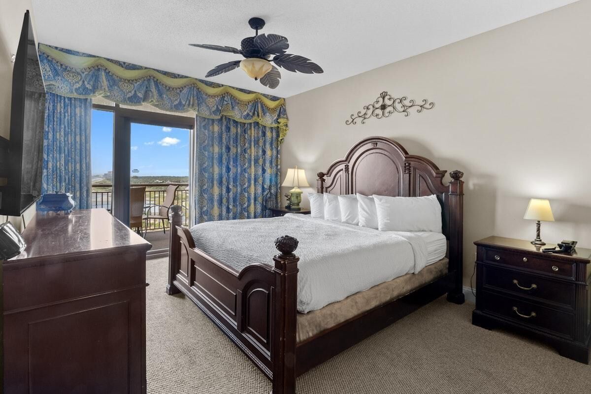 A bedroom with a large bed, two bedside tables with lamps, a ceiling fan, and a view of the outdoors through a sliding door.