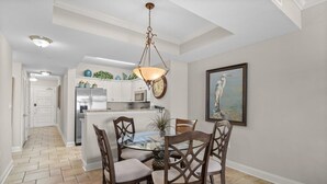 A dining area with a glass table and wooden chairs, a hanging light fixture, and a kitchen in the background.