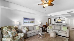 A living room with two sofas, a coffee table, and a ceiling fan. There is a kitchen in the background.