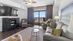 A spacious living room with a large flat-screen TV, a ceiling fan, a balcony with a view of the beach, and a comfortable seating area.