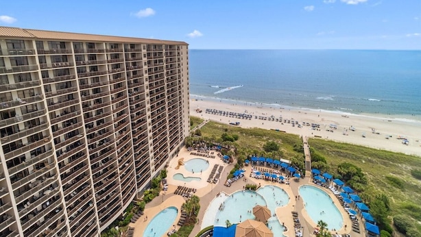 Ein Hochhaus-Hotel mit mehreren Balkonen, das auf einen Strand und einen Poolbereich mit Liegestühlen und Sonnenschirmen blickt.
