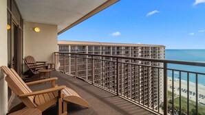 Ein Balkon mit Holzstühlen und Tisch, mit Blick auf einen Strand und ein Hochhaus.