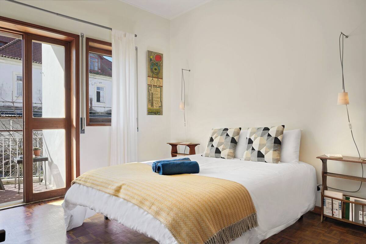 A bedroom with a large bed, a wooden floor, a small bedside table, a nightstand, and a window with curtains.