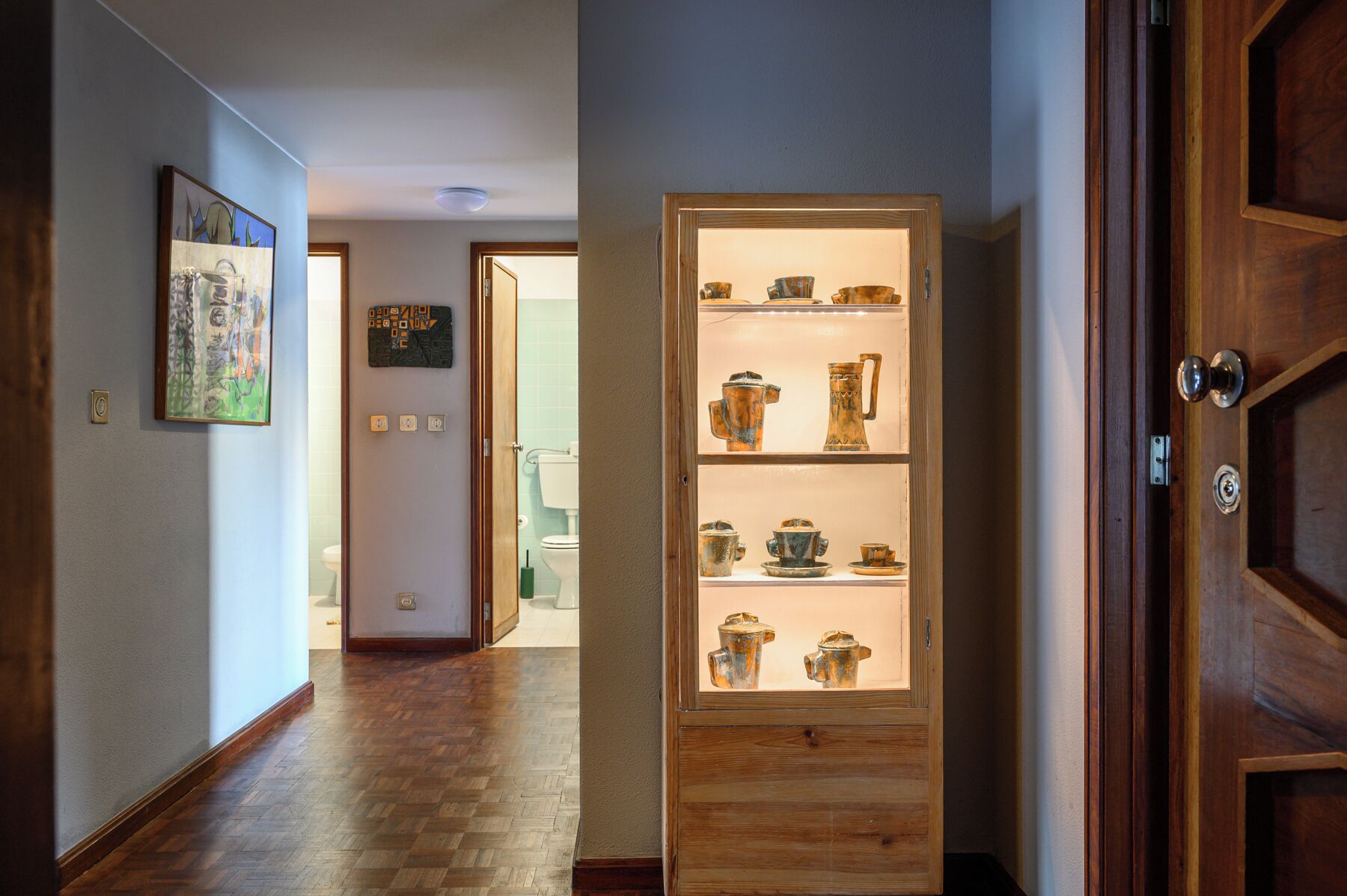 Um corredor com piso de madeira, uma vitrine de vidro com objetos de cerâmica e uma porta com moldura de madeira.