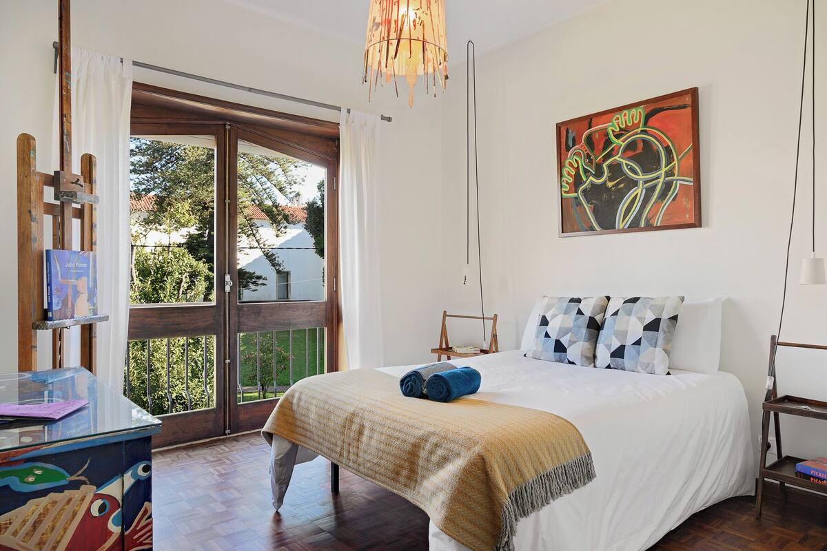 A bedroom with a bed, a painting, and a window with a view of trees and buildings.