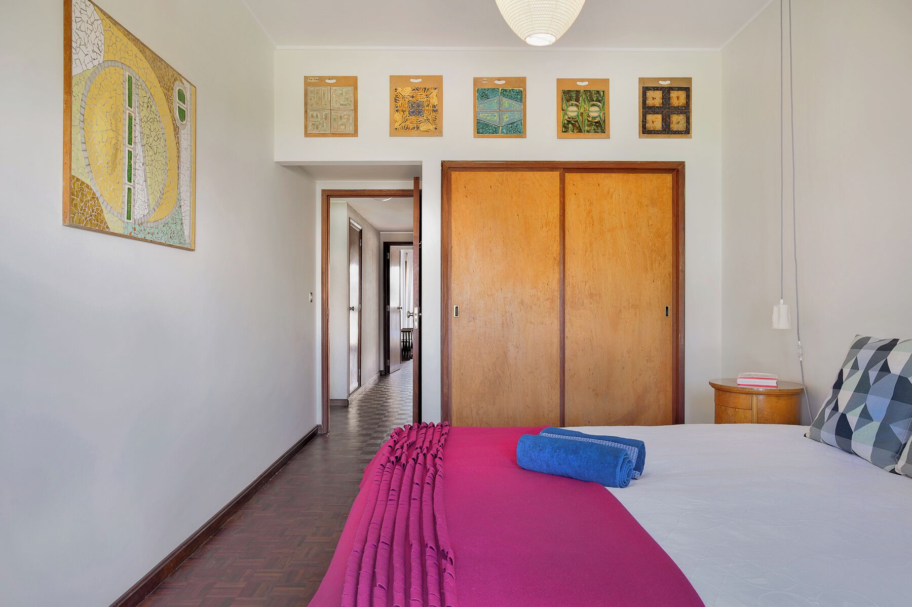 Habitación de hotel con un armario de madera, una cama con edredón blanco, una manta rosa y una toalla azul. Cuatro cuadros enmarcados decoran la pared sobre el armario.