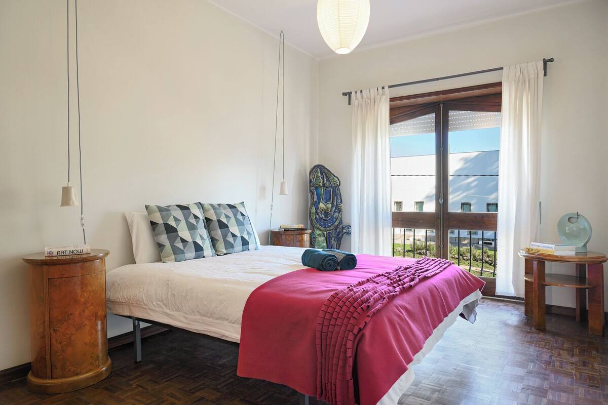 A bedroom with a bed, a wooden nightstand, a decorative mirror, and a view of the outside.