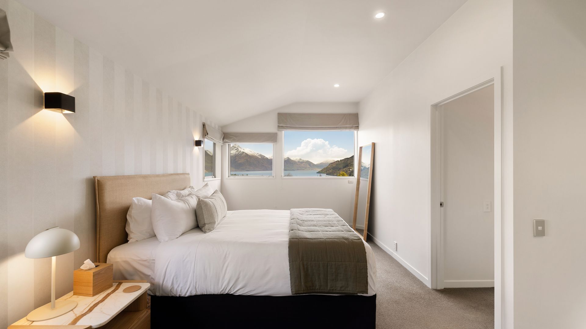 Een slaapkamer met een bed, nachtkastje, lamp en uitzicht op bergen en een meer door een raam.