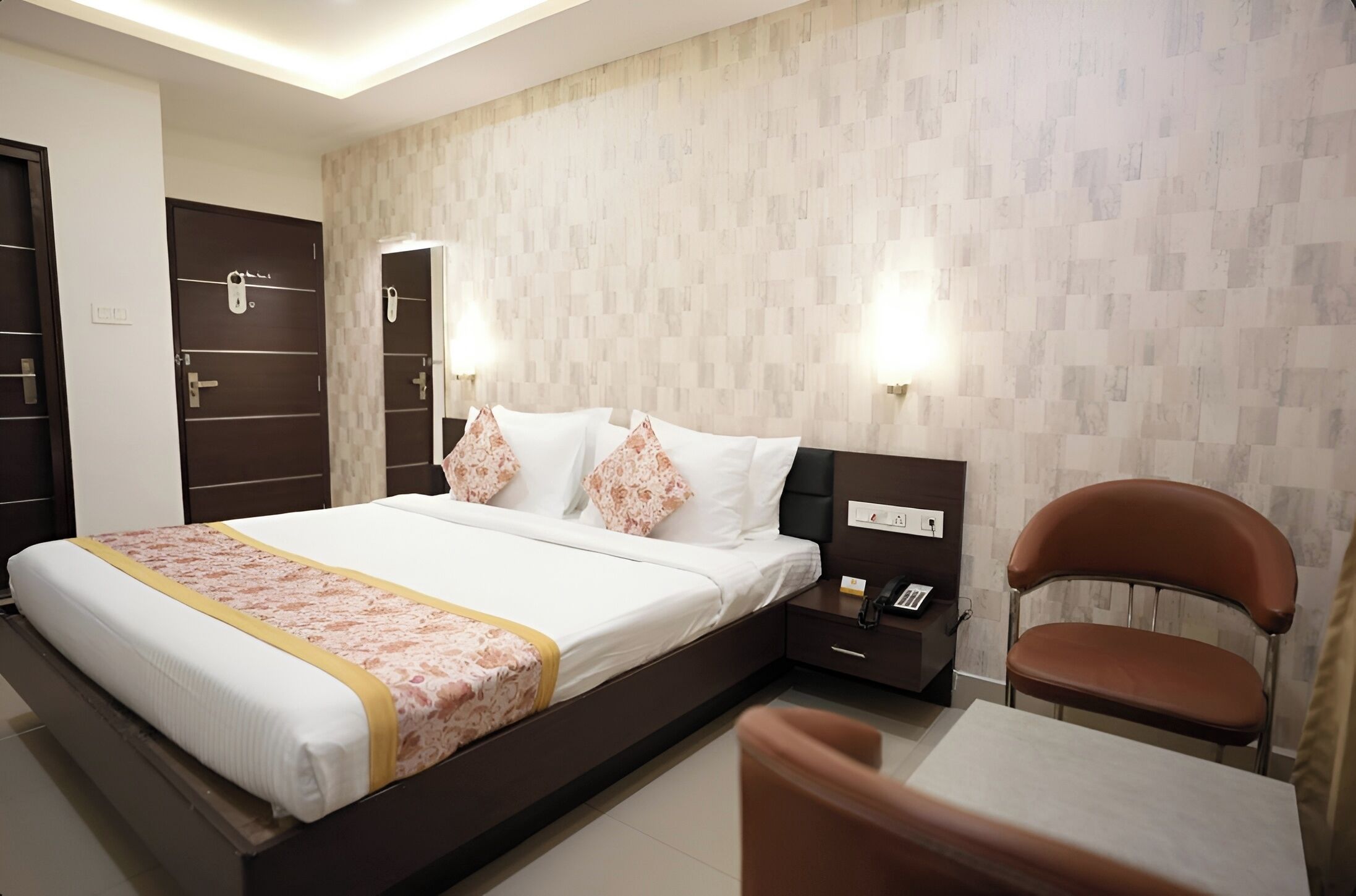 A hotel room with a bed, bedside table, chair, and a wall with a patterned design.