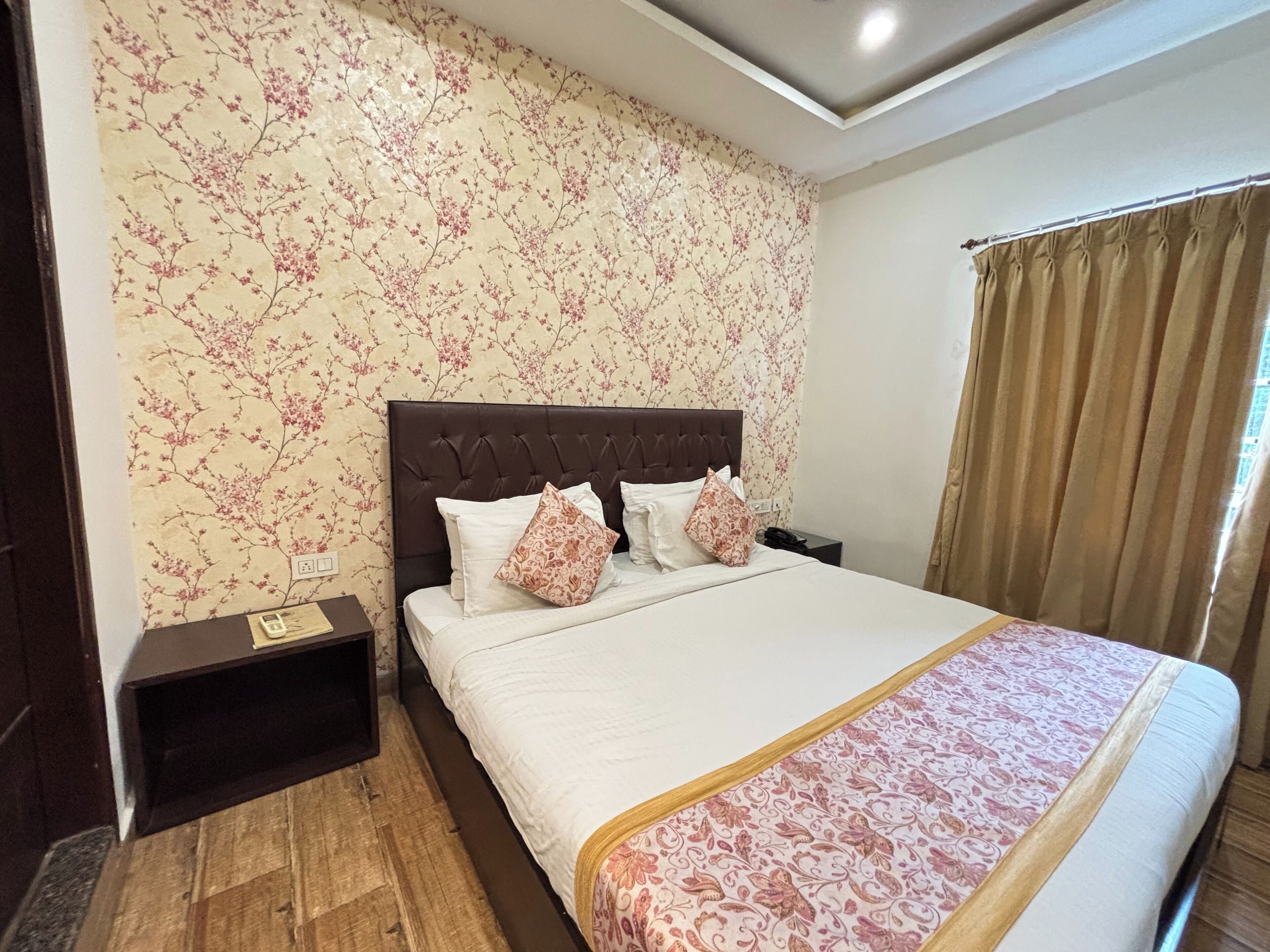 A hotel room with a large bed, a wooden nightstand, a floral wallpaper, and a window with curtains.