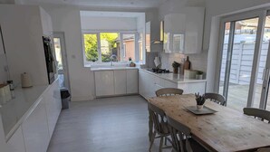 A modern kitchen with white cabinetry, a wooden dining table, and a large window providing natural light. - Cosy Bee  (Bournemouth)