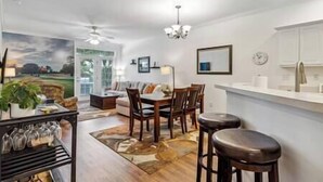 A modern kitchen with a white countertop, wooden chairs, and a dining table. There is a large window with a view of the outdoors. - Birdies & Beaches Getaway, Golf and Beach Retreat (Pender County)