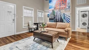 A living room with a beige sofa, a coffee table, and a painting of a street scene. There is a door and a window with blinds. - Water Street Hideaway (Wilmington)