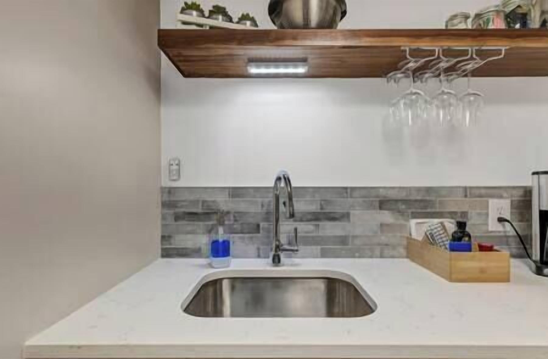 A kitchen with a stainless steel sink, a wooden shelf above, and a tiled backsplash.