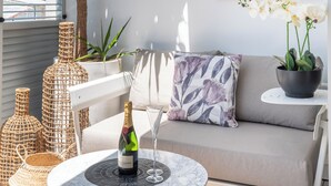 Une pièce baignée de lumière, avec un canapé blanc, une table ronde sur laquelle se trouvent une bouteille et un verre, des vases en osier et une orchidée en pot.